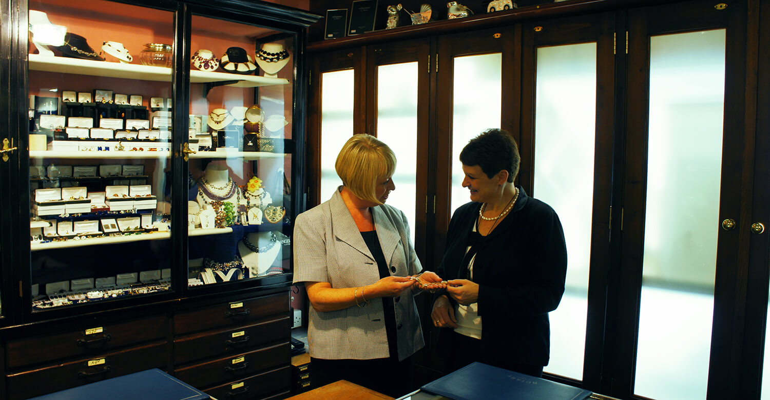 2 Angelo Smith staff members hold a piece of jewellery and smile at each other.