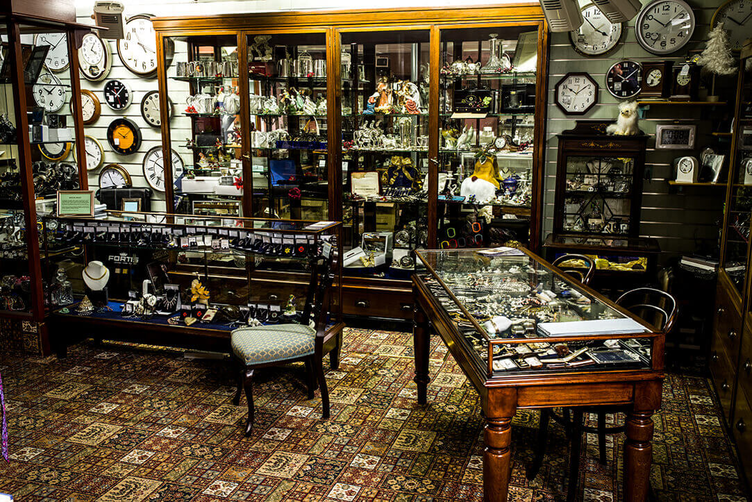 The showroom interior with two desks filled with jewellery, clocks hanging on the wall, and an empty chair in front.
